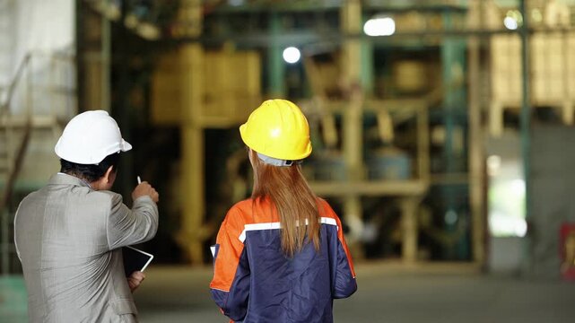 Rear view of a rice mill owner in a suit and a female engineer with long hair in hard hats, he points at the milling machinery with a pen as they walk toward the line