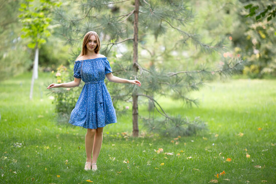 A young smiling woman strolls through a spring park in a good mood. A beautiful young girl with long light brown hair in a dress poses among plants, trees, and flowers.