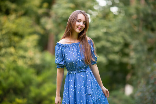 A young smiling woman strolls through a spring park in a good mood. A beautiful young girl with long light brown hair in a dress poses among plants, trees, and flowers.