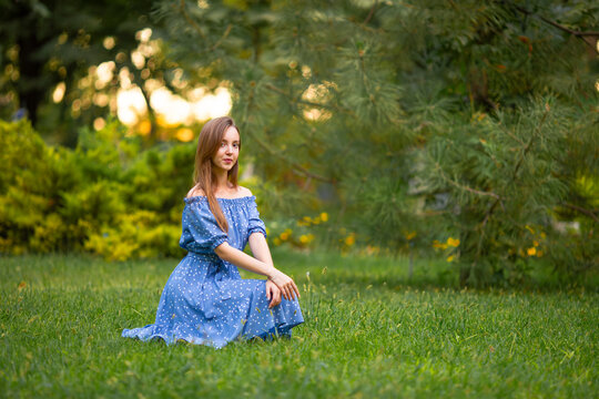 Beautiful young woman in a blue summer dress relaxing on green grass in a park, enjoying nature, sunshine and peaceful outdoor lifestyle.