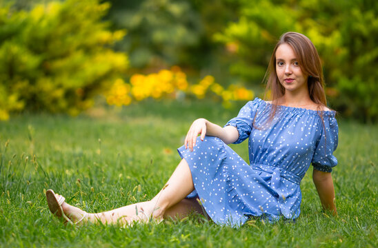 Beautiful young woman in a blue summer dress relaxing on green grass in a park, enjoying nature, sunshine and peaceful outdoor lifestyle.
