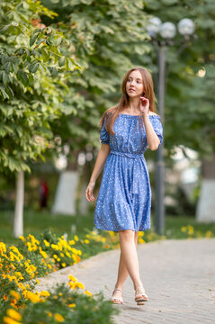A young smiling woman strolls through a spring park in a good mood. A beautiful young girl with long light brown hair in a dress poses among plants, trees, and flowers.