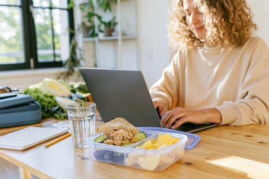 Healthy lunch with a sandwich and fruit in a lunchbox on a wooden desk, featuring a woman typing on a laptop for work life balance