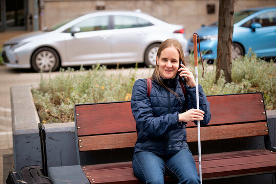 Visually impaired woman with white cane sitting on a bench and using a phone