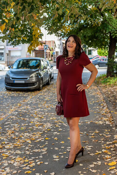 Elegant Woman in short purple dress and high heels, wearing beads necklace, carrying a small purple hand bag walking on a autumn street full of fallen leaves
