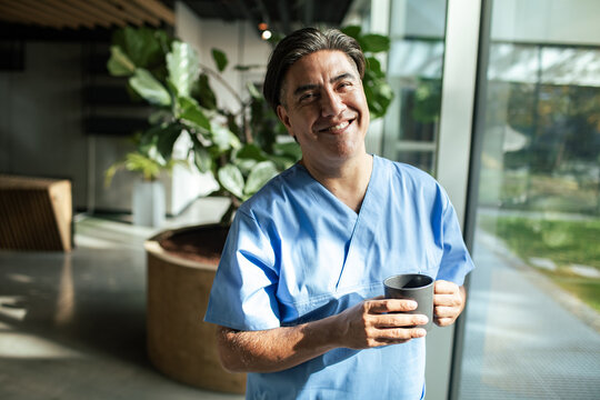 Smiling healthcare worker in scrubs holding coffee by window