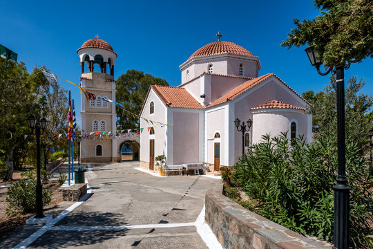Picturesque Agios Sozon Church with its distinct pink facade, terracotta-tiled domes, and separate bell tower stands under a bright blue sky in the coastal village of Perdika on Aegina Island, Greece