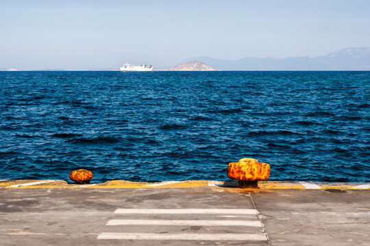 Two weather-worn, yellow mooring bollards sit along a concrete port pier overlooking deep blue sea waters with a distant passenger ferry navigating past islands near Aegina, Greece.