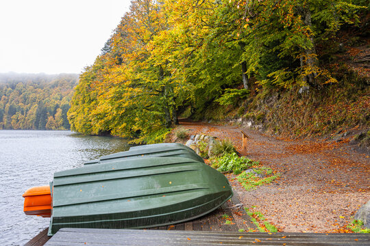 Green rowing boats rest on the shore of Lac Pavin beside a wooden dock and golden autumn forest in Auvergne. An off season travel concept evokes lake leisure, fishing and boat rental.
