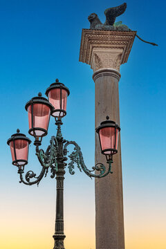 Architectural details in St. Mark&rsquo;s Square, Venice, Italy, featuring the Winged Lion of Saint Mark atop a granite column and a classic Venetian lantern with distinctive red shades.
