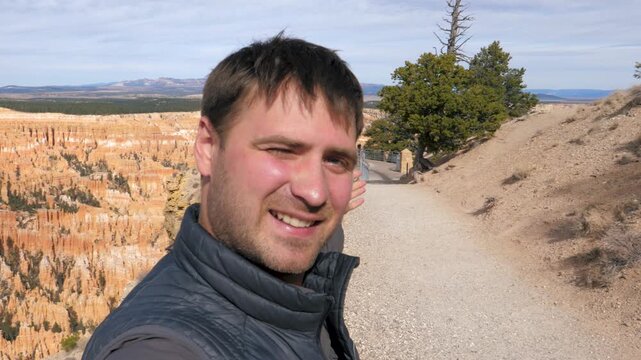Cheerful male traveler recording selfie video while hiking along scenic trail at bryce canyon national park. Man gesturing towards beautiful amphitheater orange rocks hoodoos on sunny day