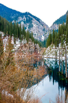 The flooded forest of Lake Kaindy in winter. A beautiful winter mountain landscape in Kazakhstan. A blue lake with tree trunks protruding from it against a backdrop of snow-capped mountains.