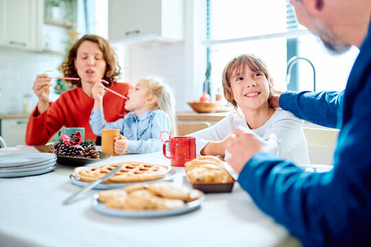 Family gathering around the kitchen table, sharing waffles and warm drinks while children play with candy canes beside festive decorations