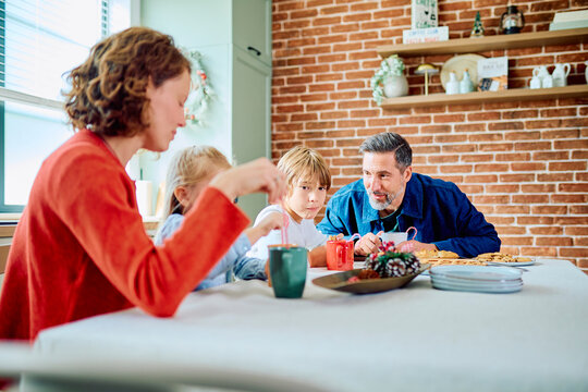 Family gathered around a kitchen table, sharing breakfast, tea and cookies in a cozy festive home decorated for Christmas