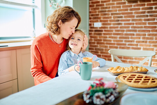 Mother kissing her smiling daughter at the kitchen table, sharing a tender holiday moment beside homemade pies and festive decorations