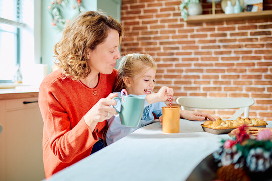 Mother and daughter sharing a cozy holiday moment, with the girl stirring hot chocolate at the kitchen table beside festive pastries