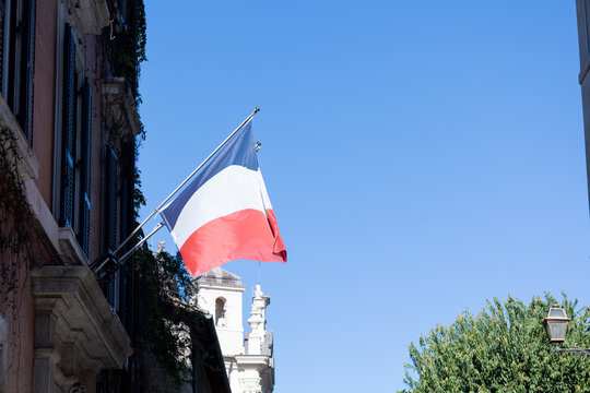 French tricolor flag hanging from a historic building facade against a blue sky in central Rome, Italy