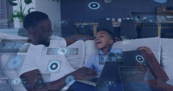 Laughing dad in white tee and son in blue shirt on sofa using laptop holographic overlays