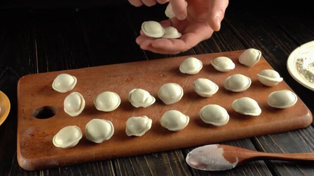 Hands gathering handmade dumplings from a wooden board beside a flour bowl and spoon during food preparation in a rustic kitchen