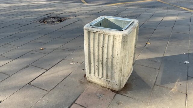 Empty concrete trash bin with metal liner stands on stone tiled pavement of city street outdoors. Urban waste management infrastructure, public sanitation service and garbage disposal container area.