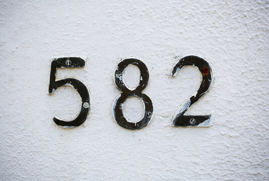 Number 582 in metal house numerals with screws mounted on whitewashed stucco wall, scratched paint, rusty screws, street address sign, residential building facade, textured plaster, urban stylin