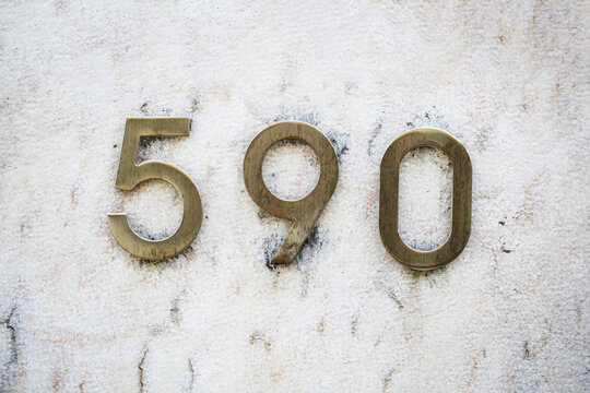 Number 590 in brushed brass house numerals mounted on weathered marble wall, stained stone surface, street address plate, metal digits, residential building facade, cracked plaster, urban styling