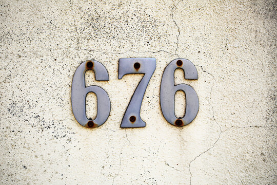 Number 676 in steel house numerals with rusty screws mounted on cracked cream stucco wall, weathered plaster surface, street address sign, metal digits, residential building facade, urban styling