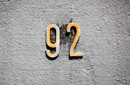92 street number mounted on a rough stucco wall exterior, vintage metal numerals indicating residential building address identification, urban architectural facade details, outdoor close-up view.