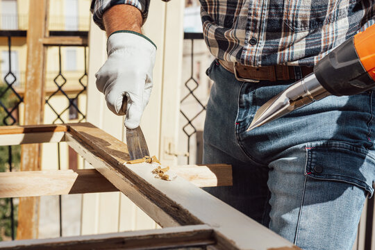 Adult carpenter wearing protective leather gloves, using a heat gun and a wood scraper to remove paint from an old wooden window. Construction industry, carpentry. Restoration. 
