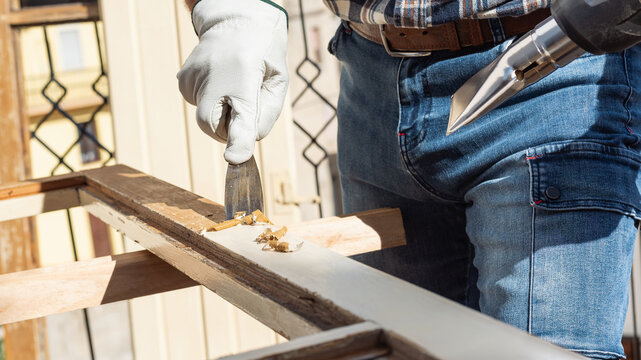 Adult carpenter wearing protective leather gloves, using a heat gun and a wood scraper to remove paint from an old wooden window. Construction industry, carpentry. Restoration. 