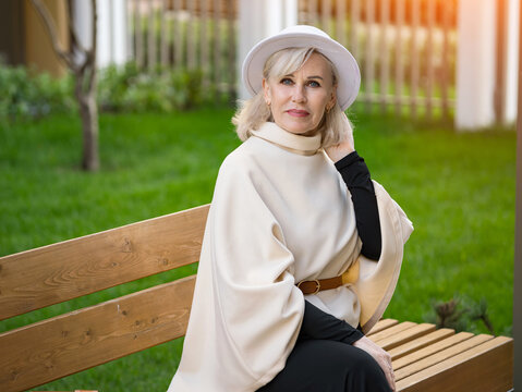 Confident older woman rests on wooden garden bench in refined white hat and cream clothing. Fashionable woman touches her hair with serene expression, highlighting polished outdoor style.