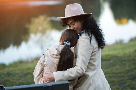 Gentle mother holds daughter close during peaceful lake contemplation in park setting. Thoughtful woman embraces young child near water, sharing emotional comfort amidst autumn scenery.