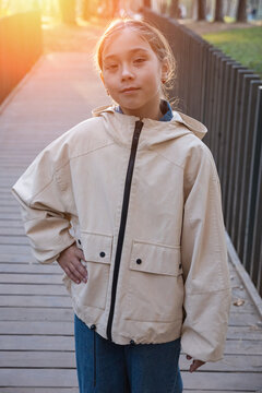 Young girl wearing beige jacket poses confidently on wooden pathway outdoors during golden hour sunset, expressing calm demeanor while standing near park fence in soft warm backlight glow.