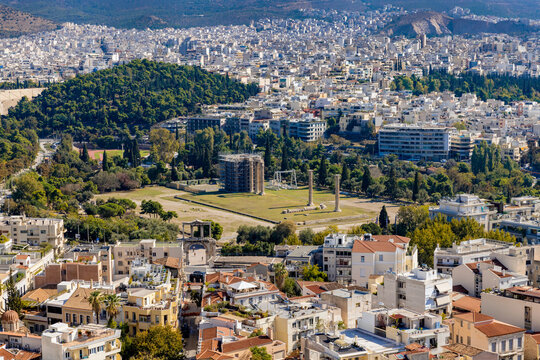 Elevated view from the Acropolis looking down at the Temple of Olympian Zeus and Hadrian's Gate surrounded by the dense urban cityscape of Athens under bright sunlight.