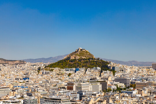 A sweeping panoramic view of Athens, Greece, looking from the Acropolis towards the prominent peak of Mount Lycabettus rising above the sprawling white urban cityscape under a clear blue sky.