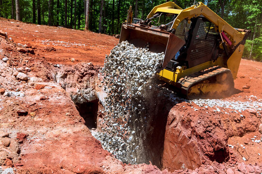Excavator machine pours gravel into trench in construction site ground is red uneven.