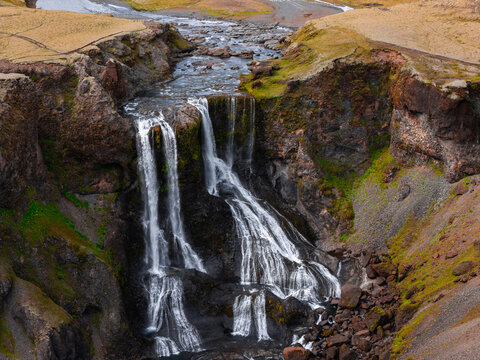 Aerial perspective captures Fagrifoss waterfall pouring over dark basalt cliffs and rocky terrain into a river gorge in the volcanic highlands of southern Iceland.