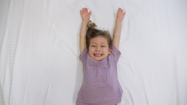 Smiling Young Girl Relaxes On White Bedding While Waving Arms Playfully. Joyful Toddler Wakes Up With Huge Smile, Stretching Body In Bright Bedroom During Wonderful Weekend Morning At Home.