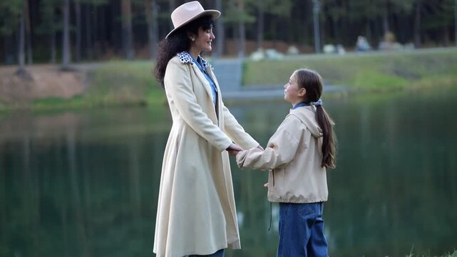 Smiling Mother Talks To Daughter Beside Scenic Woodland Lake. Stylish Woman Wearing Fedora Hat Lovingly Looks Into Eyes Of Young Girl Holding Hands Together Outdoors During Gentle Family Moment.