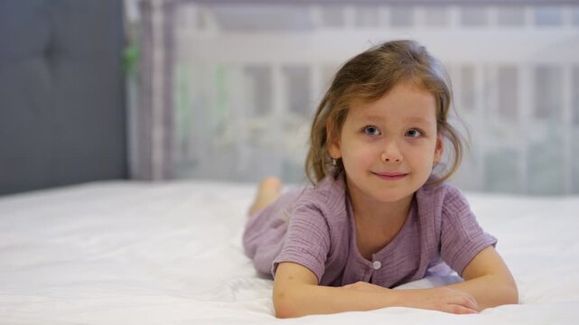 Cheerful Child Rests On Soft Mattress Displaying Bedtime Resistance, Grinning Warmly Indoors. Cute Daughter Delays Sleep Time While Relaxing Peacefully On White Bedding Inside Home Bedroom.