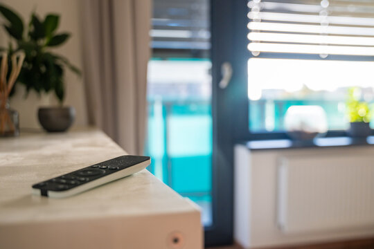 Remote control for motorized window blinds lies on a shelf with blurred blinds in the background. Smart home automation allows convenient daylight control and sun protection with selective focus
