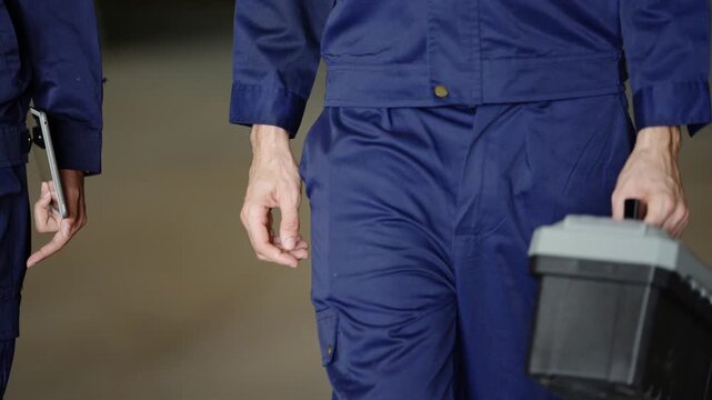 Close-up of hands carrying a grey toolbox and a tablet as rice mill workers in navy coveralls walk past camera, ending on the empty cracked concrete floor, second part