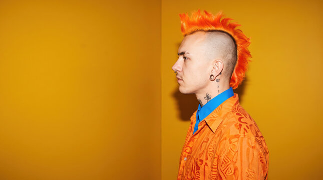 Young Man With Orange Mohawk Hairstyle Stands Against Yellow Wall in Urban Setting