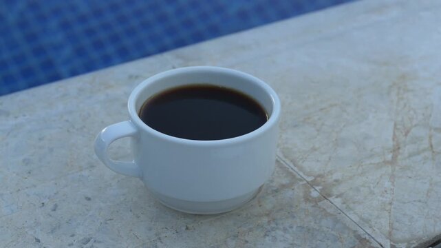 Coffee sits still in white cup beside swimming pool, creating peaceful moment for mindful pause. Poolside coffee and subdued surroundings suggest tranquility and slow living. Cup stands beside blue