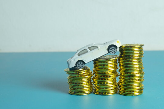 White miniature toy car climbing on ascending stacks of gold coins on a blue background. Concept of car loan, auto insurance, rising fuel price, vehicle finance, and automotive investment.