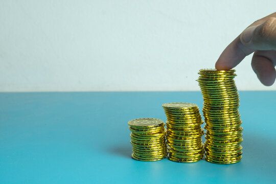 Hand placing a gold coin on ascending stacks of coins on a blue background. Concept of financial growth, business investment, wealth accumulation, and savings success with copy space.