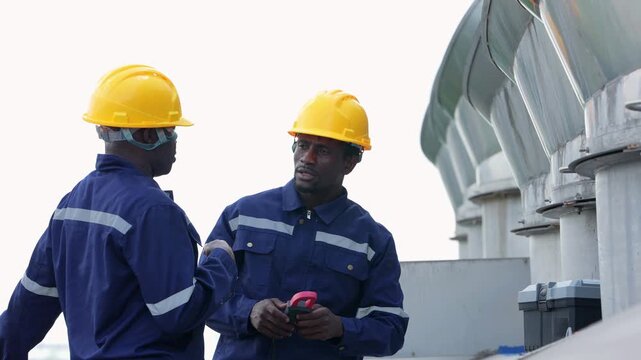 Two African engineers in yellow hard hats and navy coveralls stand on the factory roof pointing up at evaporative condenser fans while holding a clamp meter in Thailand