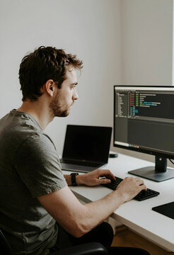 Software development and remote work, young male programmer writing code on computer at home office desk with focus and concentration on project