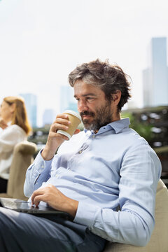 Coffee break, laptop and businessman as seated professional sips takeaway drink beside computer on terrace. Work life balance, productivity and relaxation accompany flexible working outdoors.