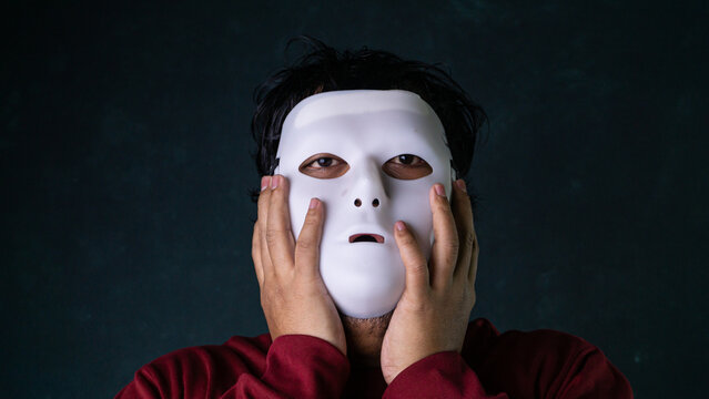A stressed Asian man wearing a blank white mask presses both hands against his artificial face under moody lighting against a dark green textured backdrop, depicting hidden shock.